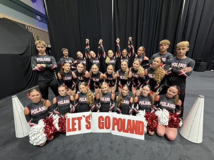 Grupa cheerleaderek z Olsztyna w strojach z napisem POLAND, trzymająca transparent z napisem LET'S GO POLAND, na tle czarnej kurtyny.