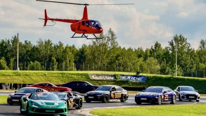 Helikopter nad grupą sportowych samochod&oacute;w na torze wyścigowym, w tle las i niebo z chmurami.