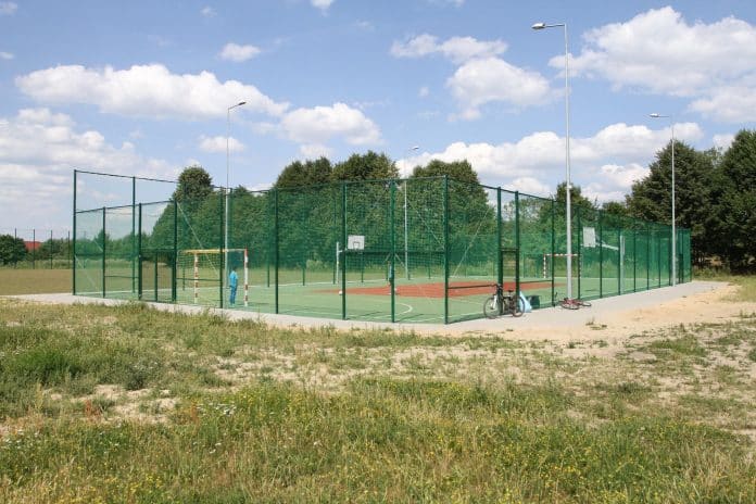 Boisko sportowe z siatką, koszami i bramkami na tle zielonych drzew i nieba z chmurami.